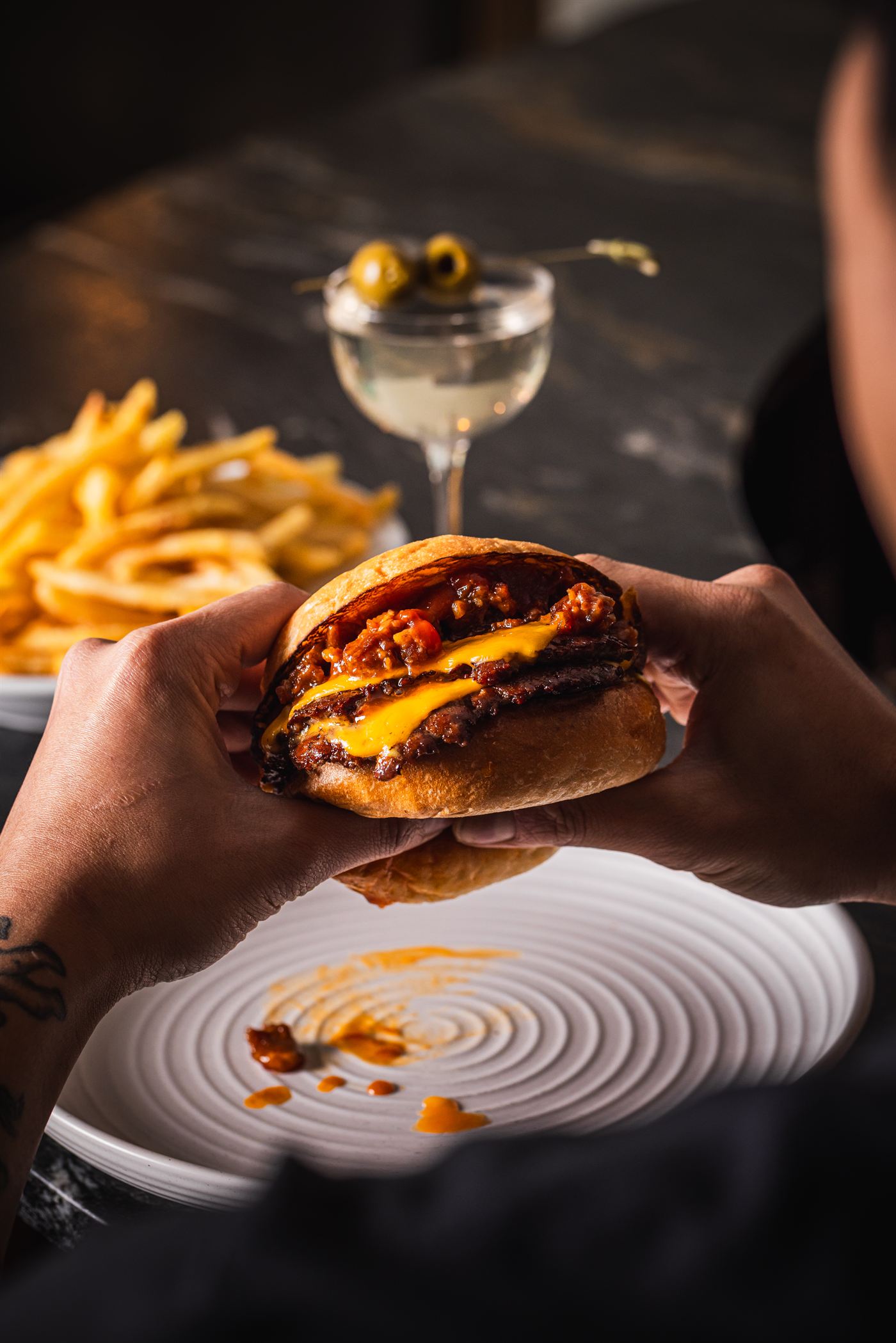 Hands holding the Burger Victoria, dirty martini and shoestring fries behind