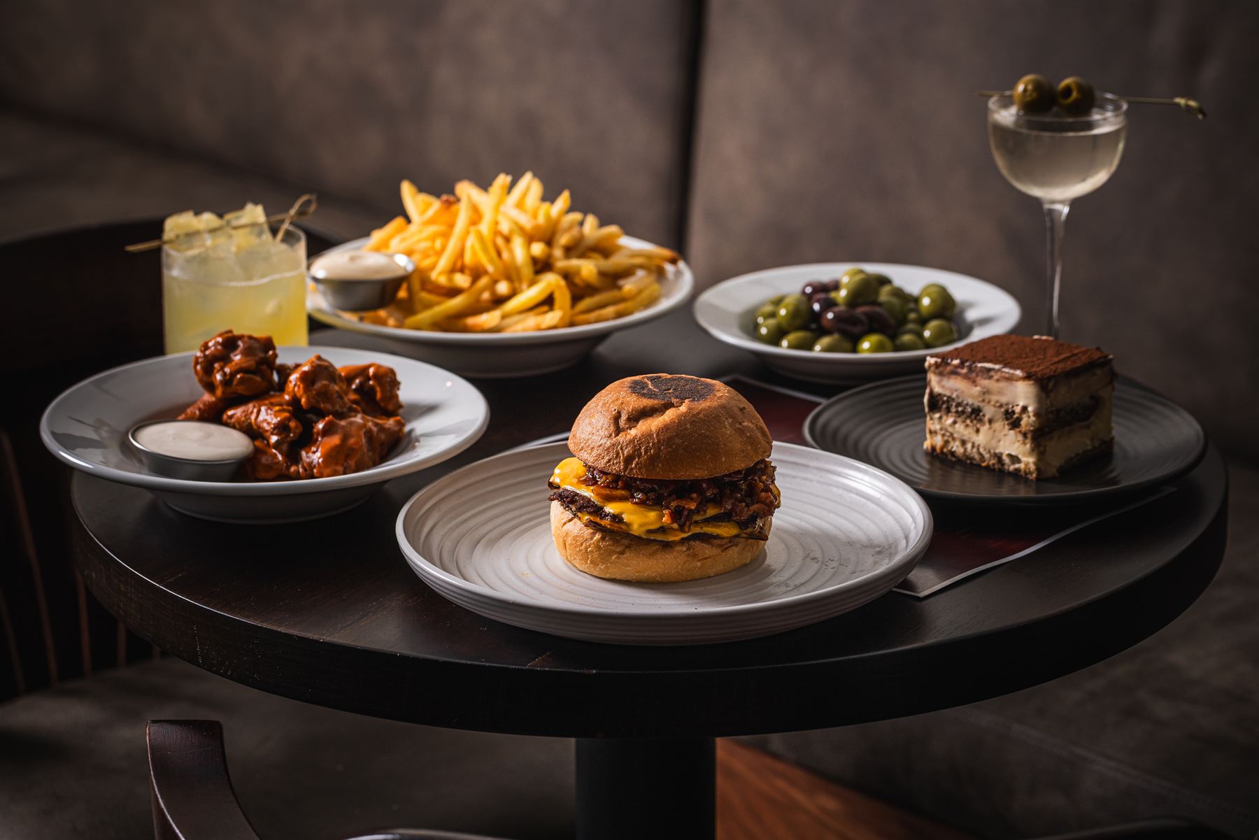 The Bar Victoria table — Burger Victoria, shoestring fries, buffalo wings, olives, tiramisu and a dirty martini on a dark timber table